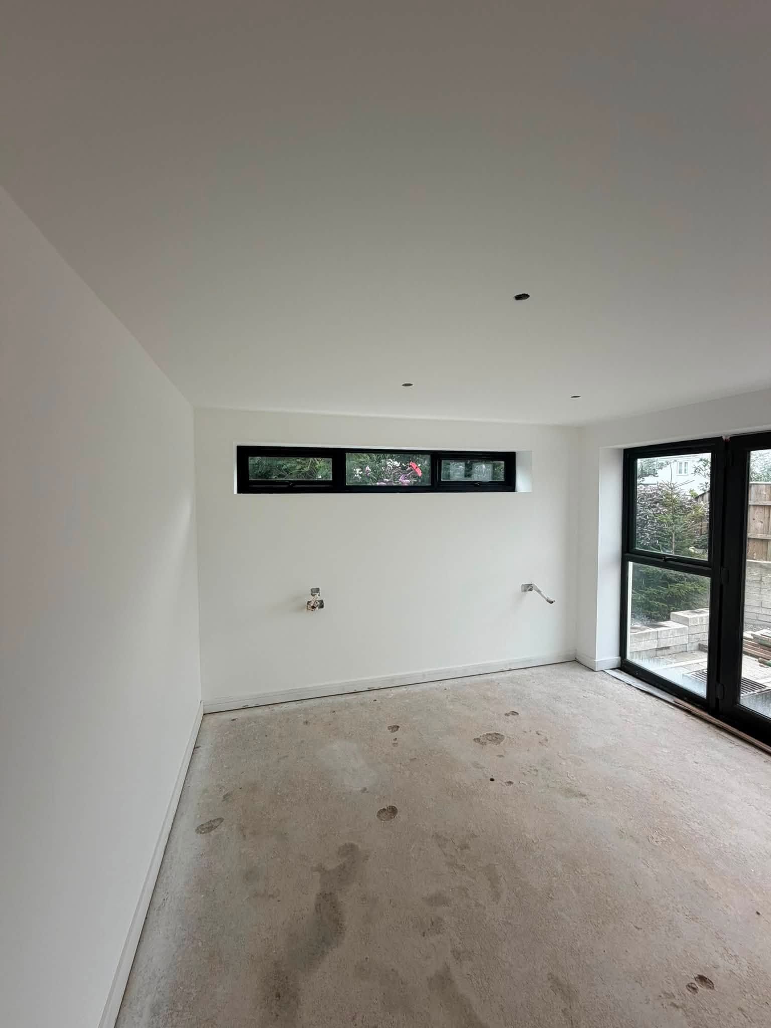 Fresh white paint after skimming in a room with black-framed glazing and an unfinished floor
