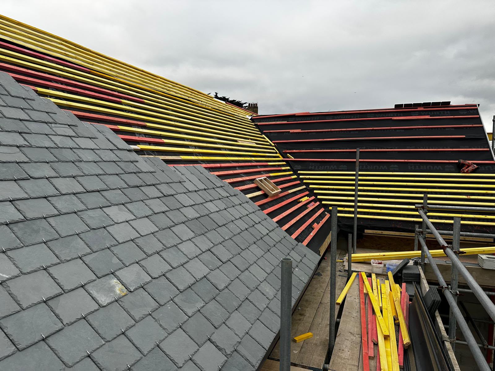 Roof with slate installation in progress beside exposed red and yellow battens and scaffolding